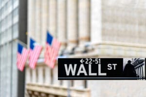 Wall Street sign with blurred American flags and building columns in the background.